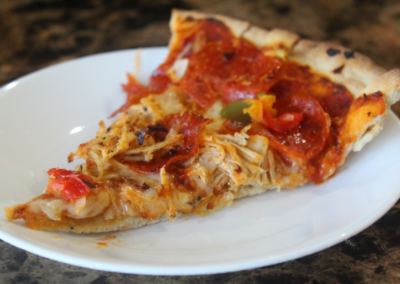 slice of homemade thin-crust cast iron skillet pizza with pepperoni bell peppers and shredded chicken facing left on white plate on granite countertop