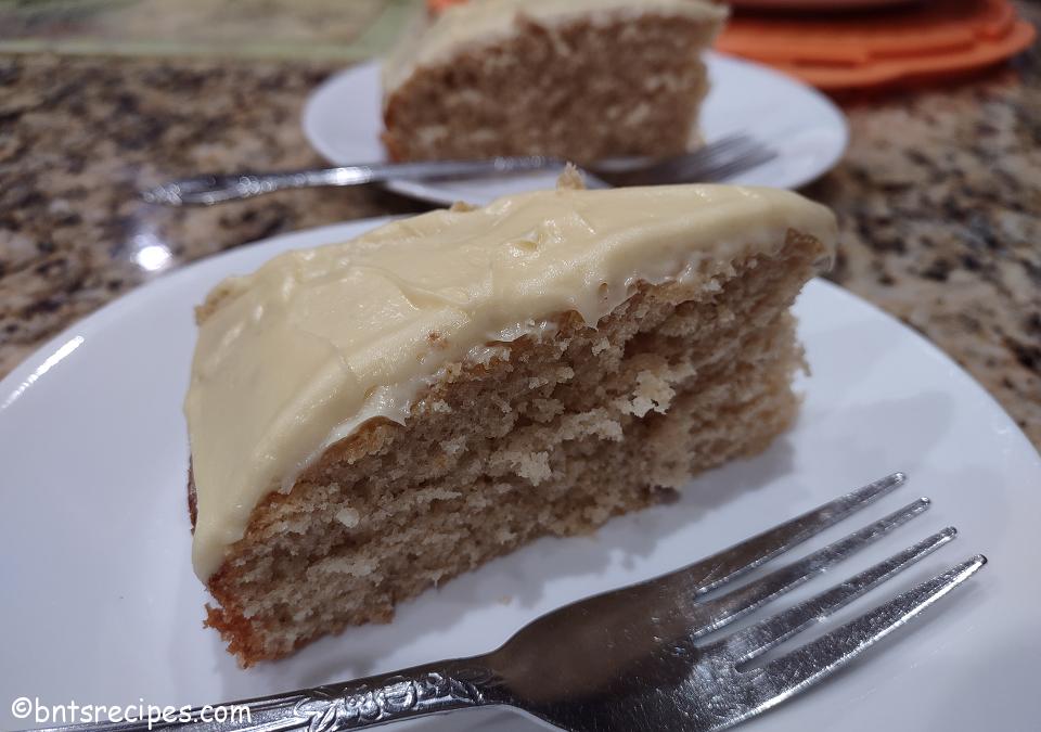 close-up of two slices of homemade grown-up yellow cake with buttercream frosting on white plates and orange cake pan on top of a granite countertop