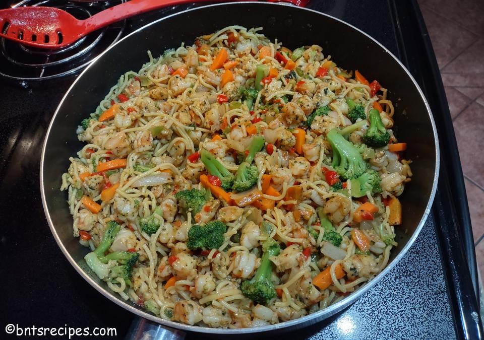 large pan of shrimp stir-fry with noodles and vegetables on black stove with red slotted spoon on hot plate nearby